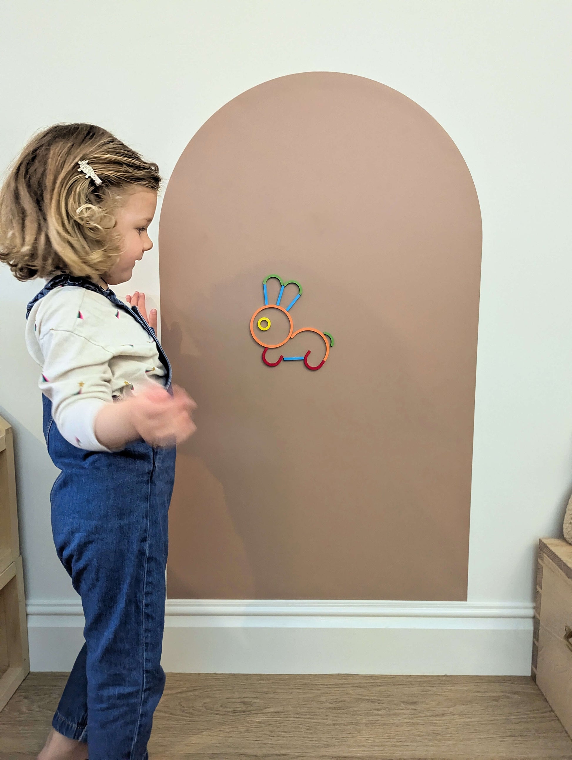 a child looking at a colourful rabbit made out of colourful wooden shapes on a brown board